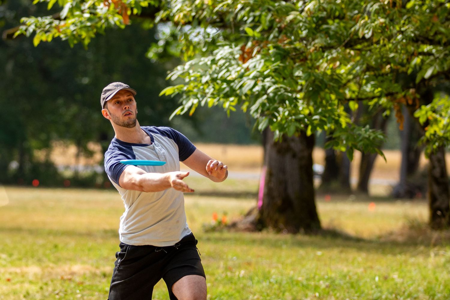 Man in white shirt throwing a disc golf frisbee in a sunny open park with trees.