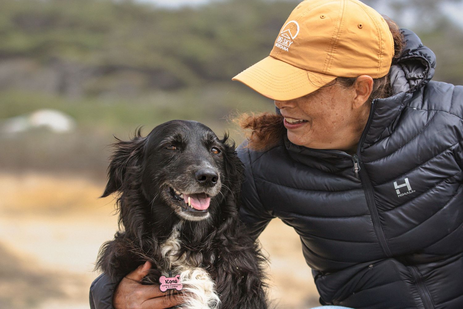Woman in orange cap hugging black dog outdoors.