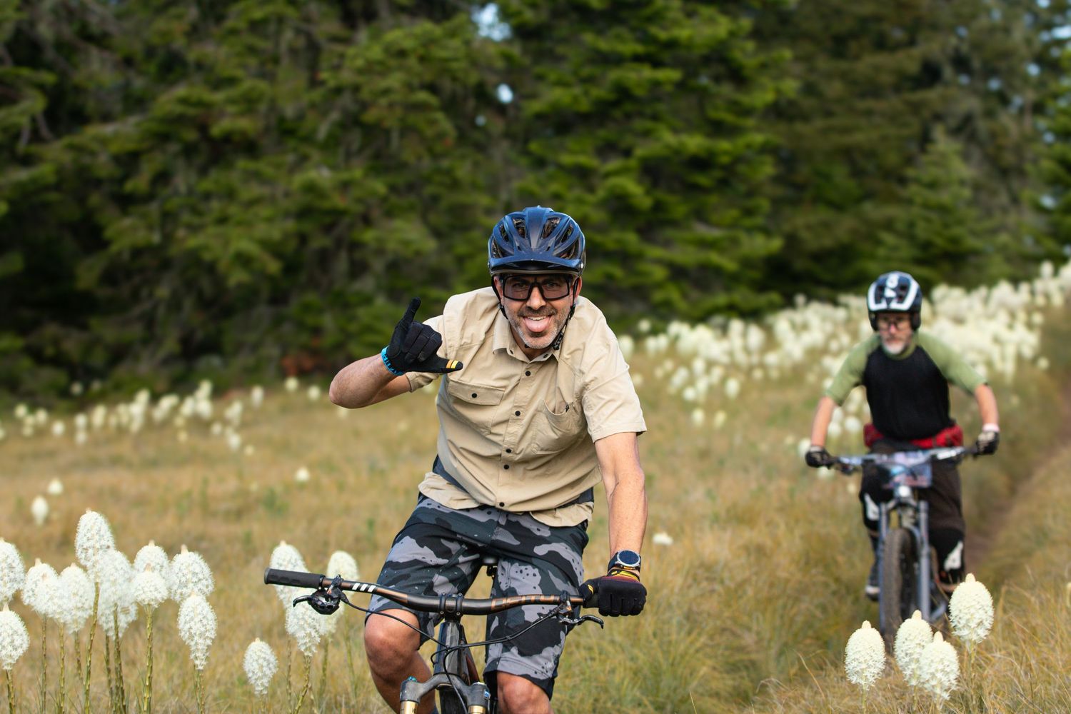 Two mountain bikers riding through field of white wildflowers with forest backdrop.