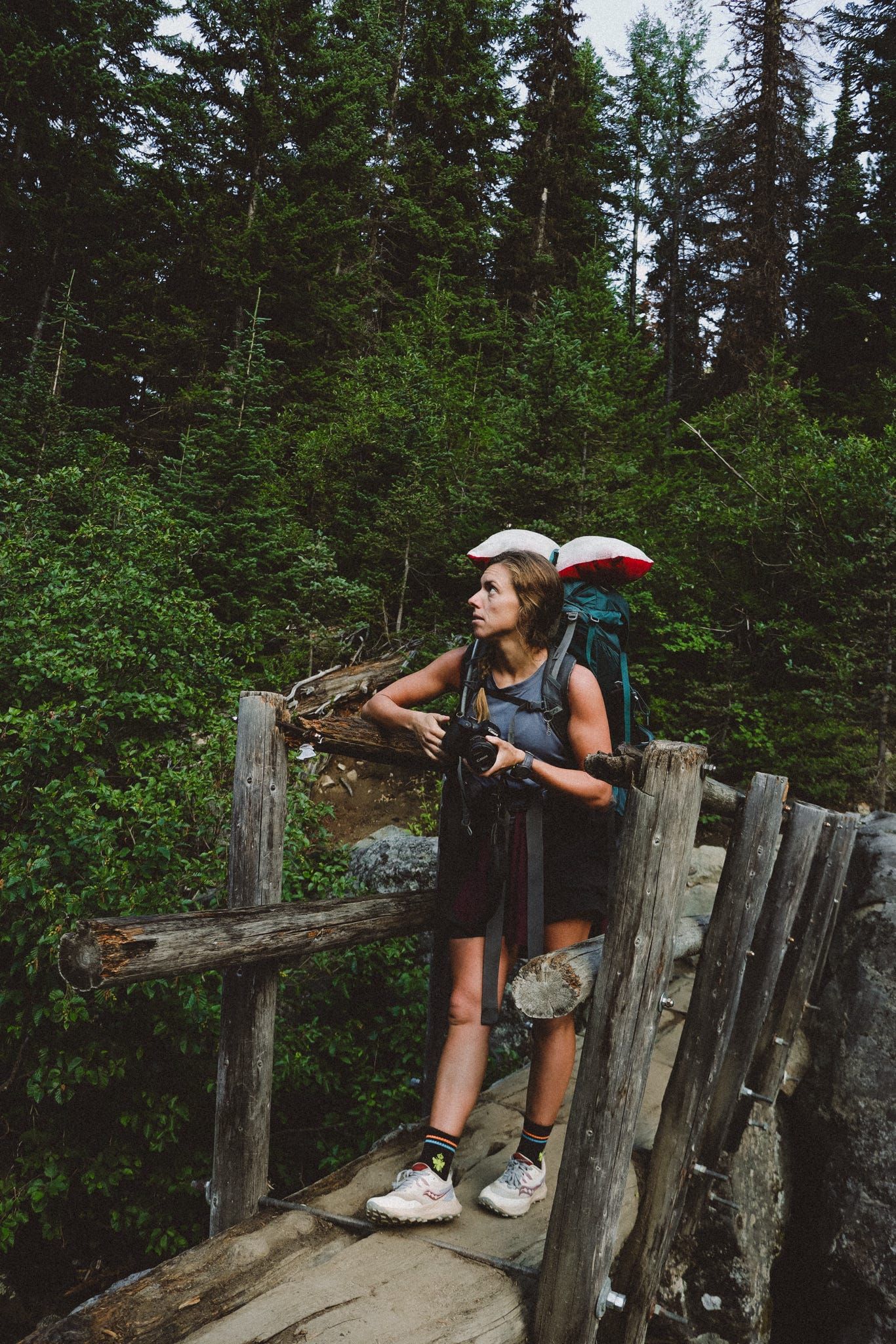 Female hiker with large backpack standing on rustic wooden bridge surrounded by lush forest.