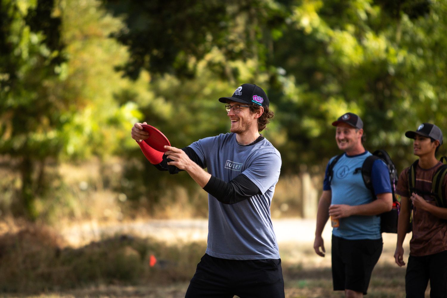 A smiling man prepares to throw a red disc golf frisbee outdoors with friends watching behind him.