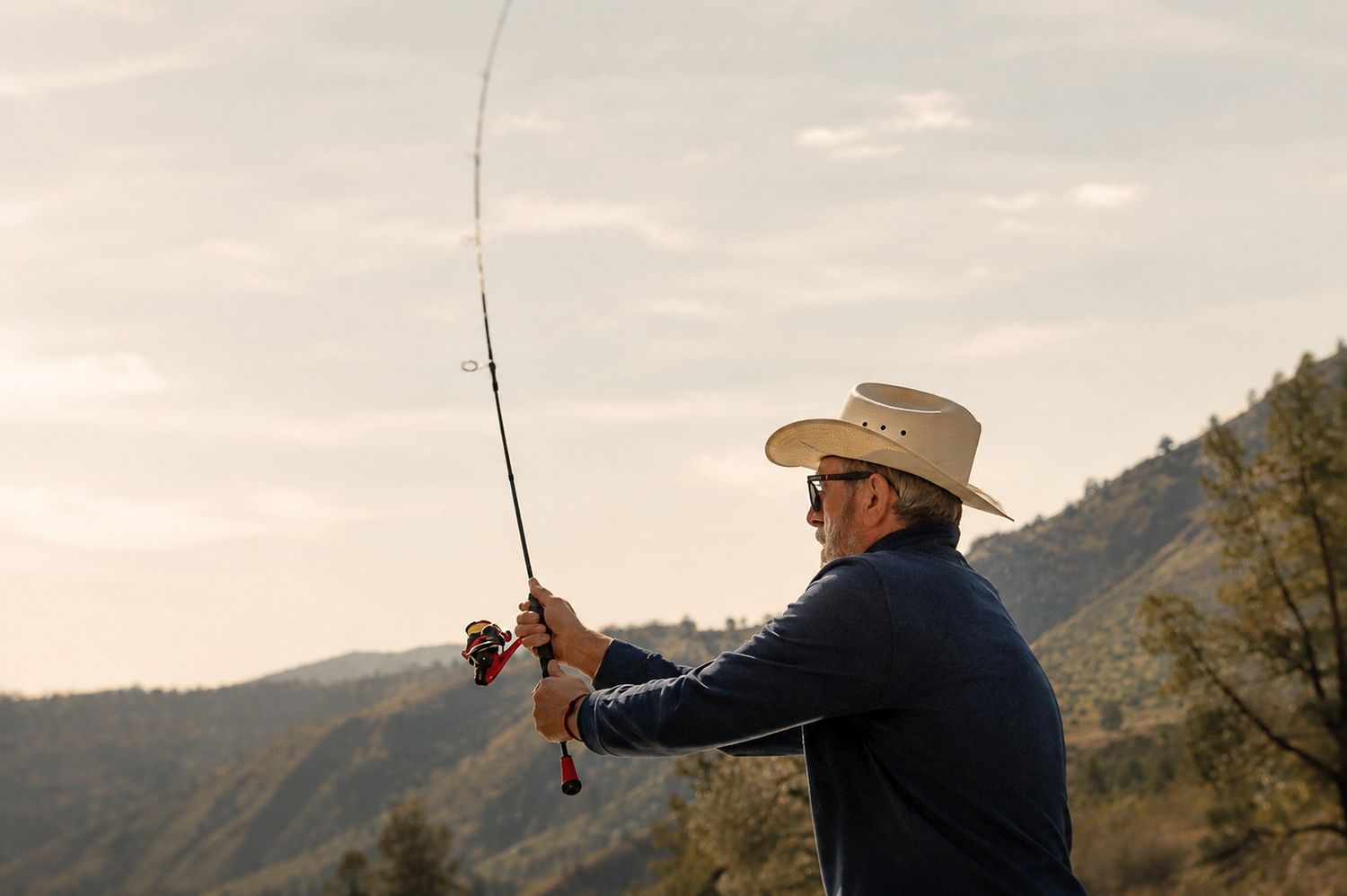 A man in a cowboy hat casts a fishing rod against a mountain backdrop at dusk.