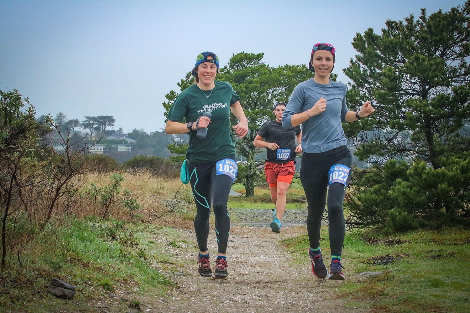 Two women wearing race bibs run together on a dirt trail through a misty landscape.