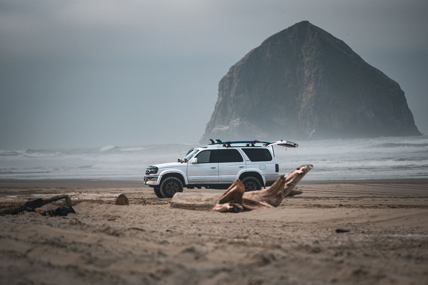 White SUV parked on a sandy beach with a dramatic sea stack rock formation rising from the misty ocean.