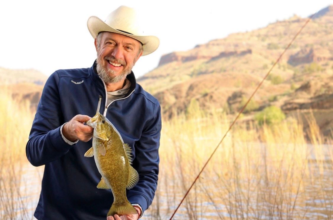Smiling man in cowboy hat holding a freshly caught bass fish while fishing at a scenic lake.