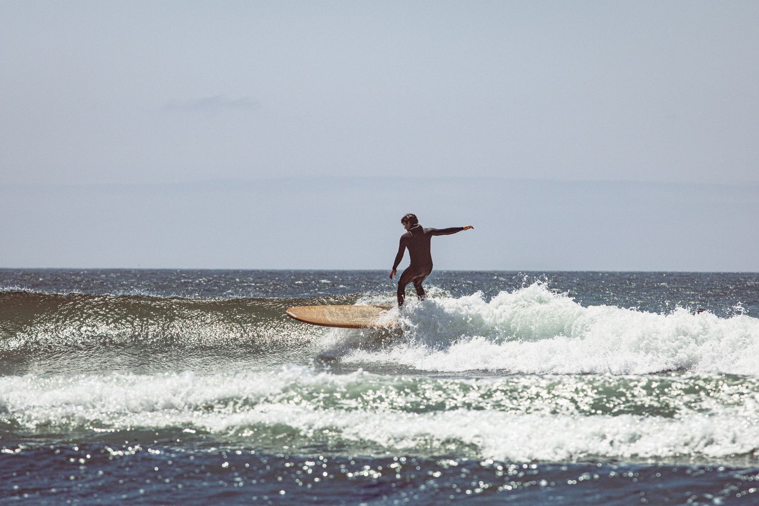A surfer rides a longboard on a rolling wave with arms outstretched under a clear sky.