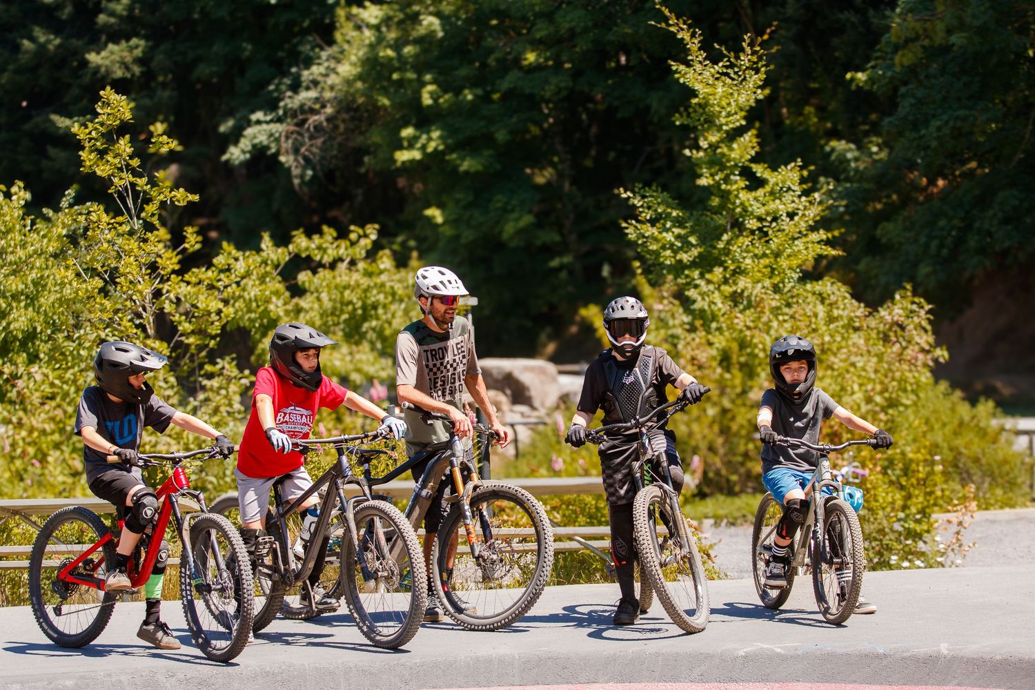 Five children wearing helmets pose together on mountain bikes on a sunny paved path surrounded by trees.