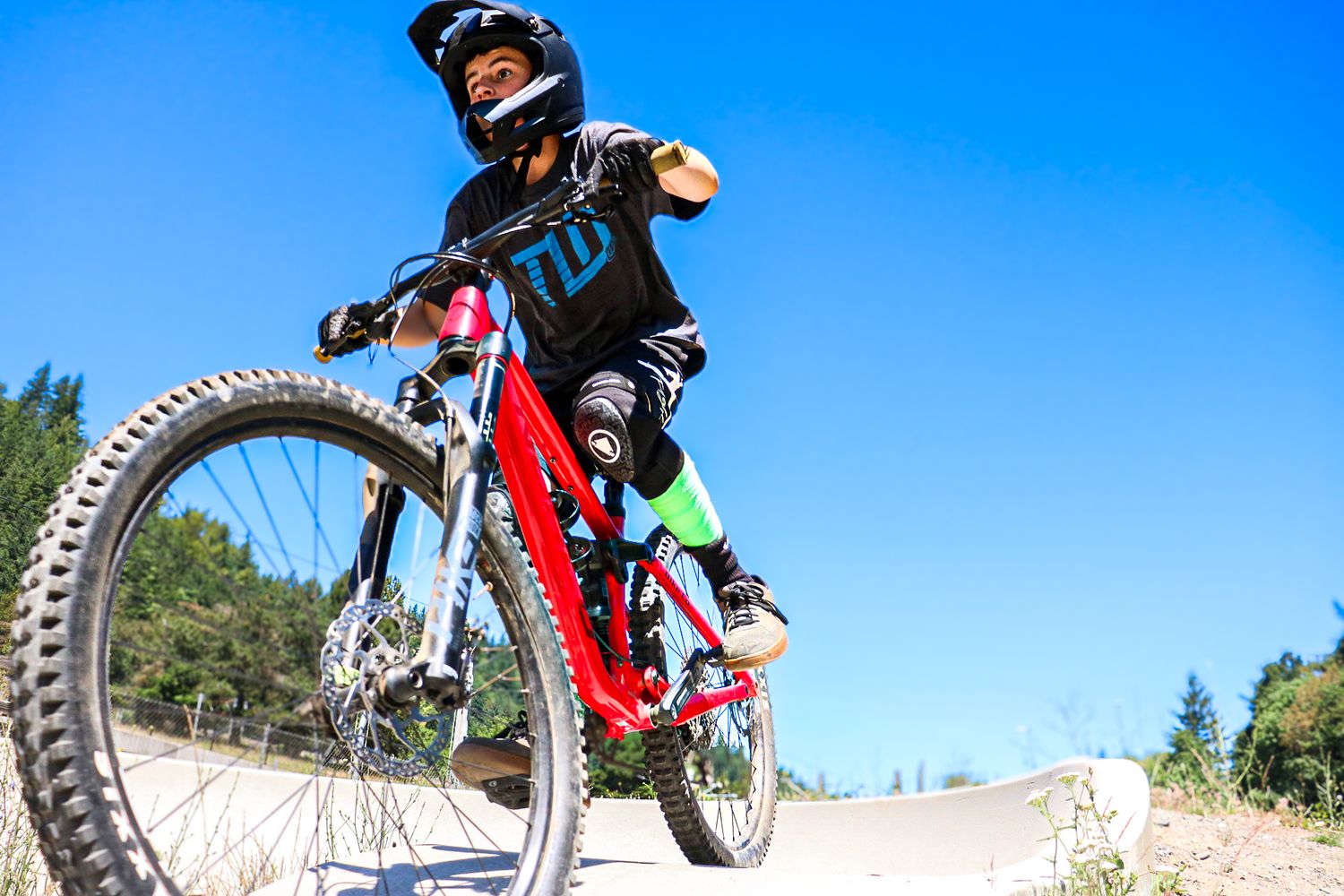 A young rider in full protective gear poses on a red mountain bike at an outdoor bike park.