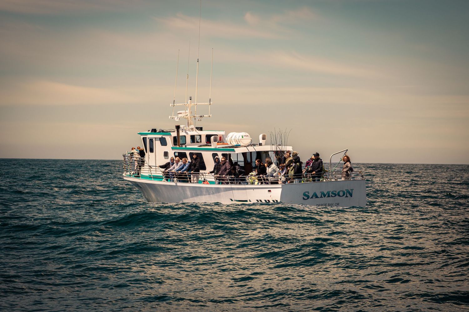A white charter boat named Samson carries passengers across calm ocean waters under a cloudy sky.
