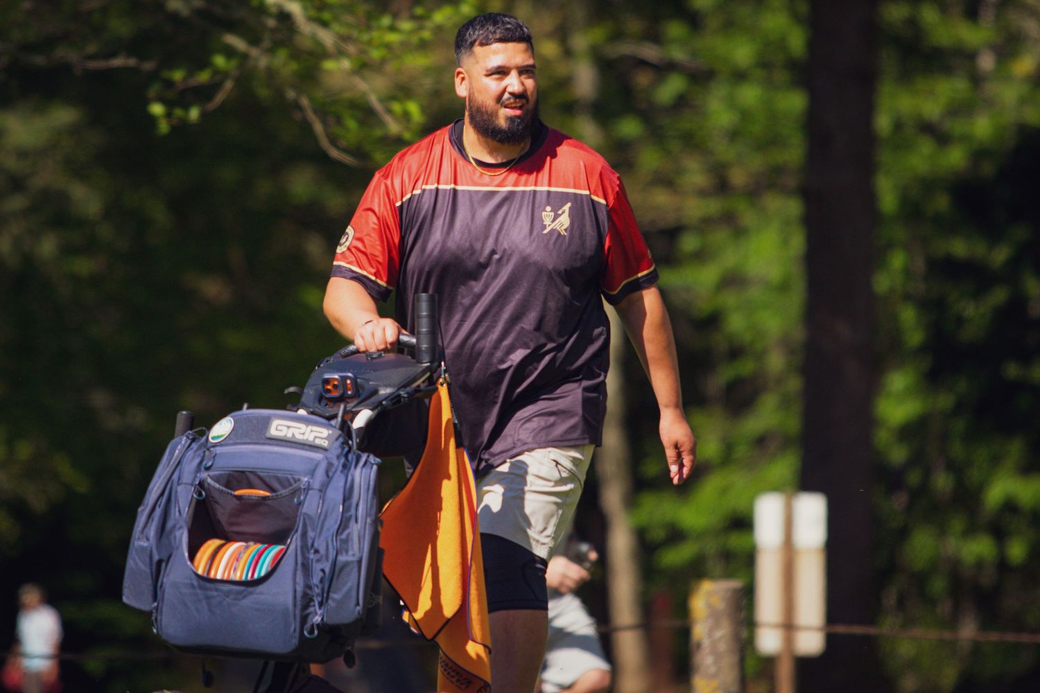 Large man carrying heavy sports bags walking along a tree-lined path outdoors.