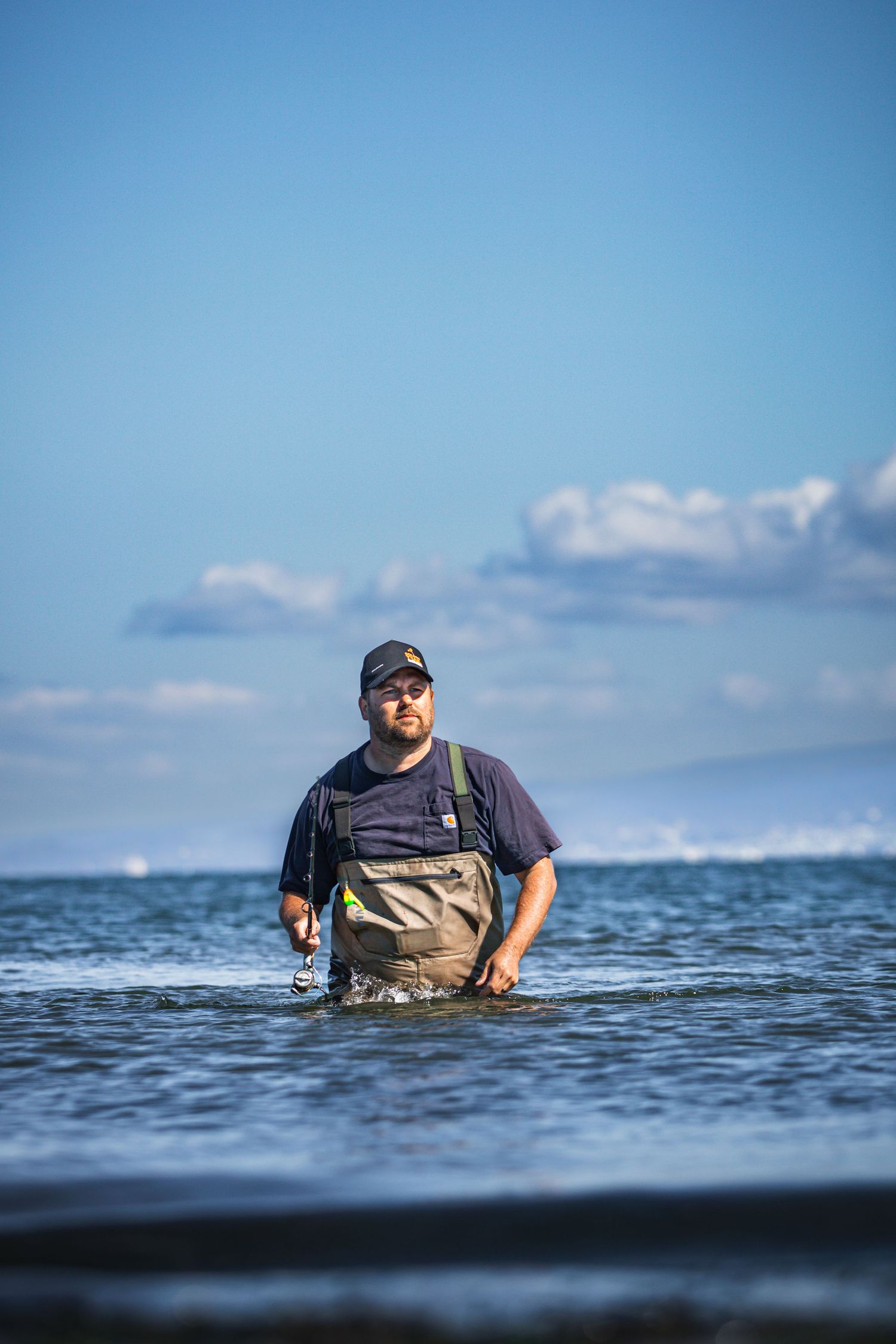 Man in waders and cap stands waist-deep in ocean water while fishing on a sunny day with blue skies.