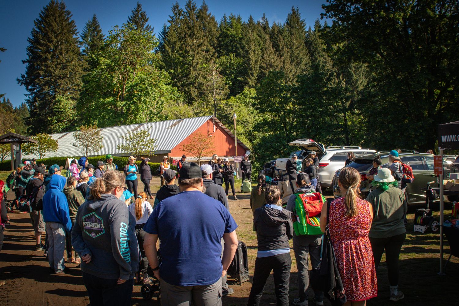 Large crowd of people gathered outdoors near a red barn surrounded by tall evergreen trees.