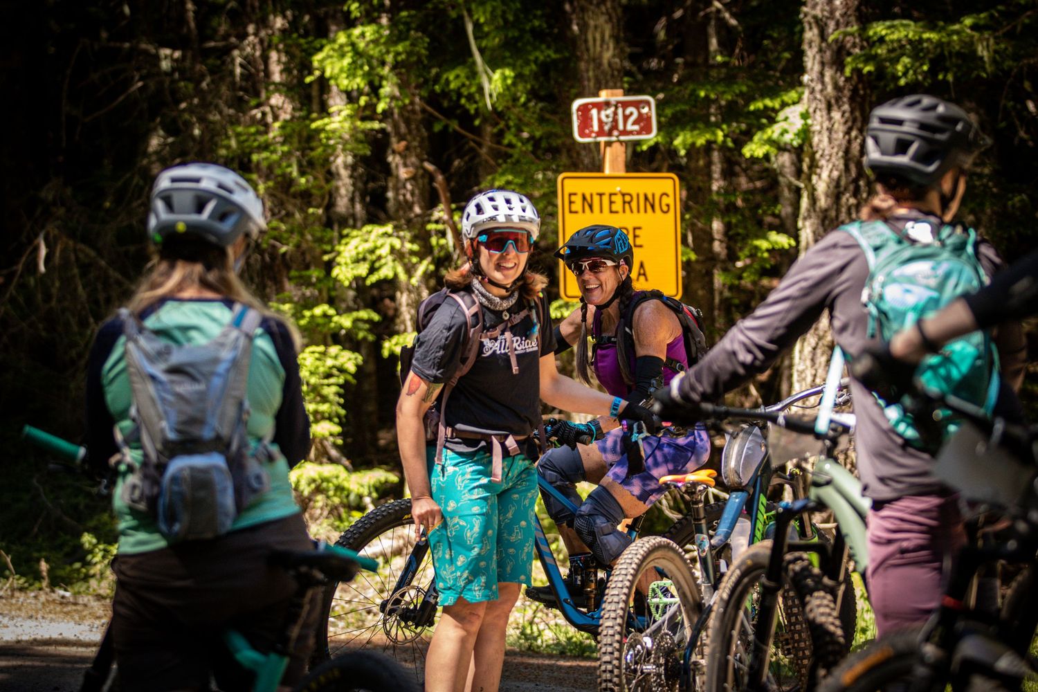 Group of female mountain bikers pausing at an entering town sign in a forest.