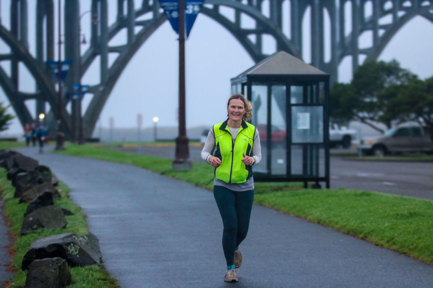 Woman in neon yellow vest jogging on a path near a large steel bridge on a foggy day.