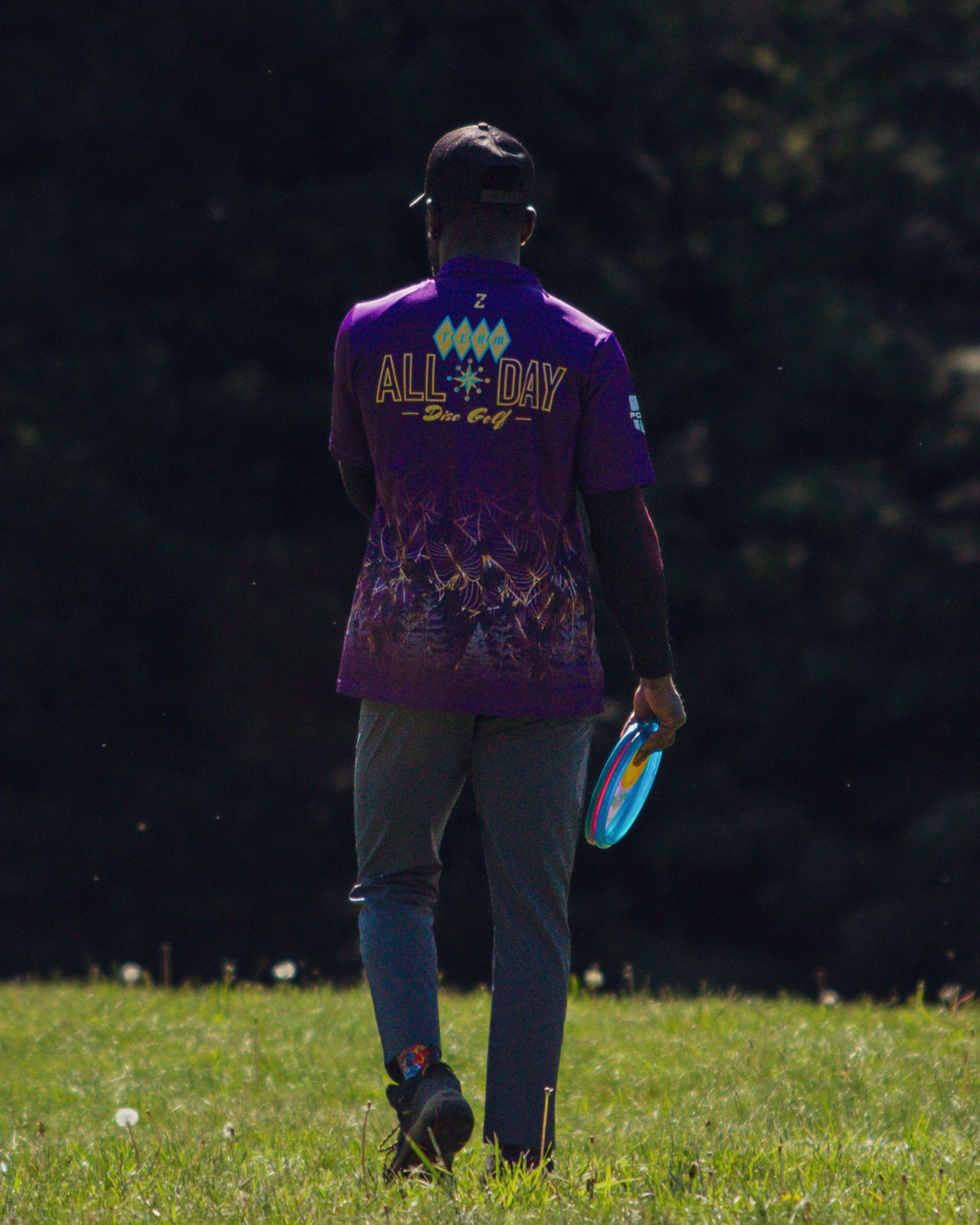A disc golfer wearing a purple All Day Disc Golf jersey walking across a sunny green field.