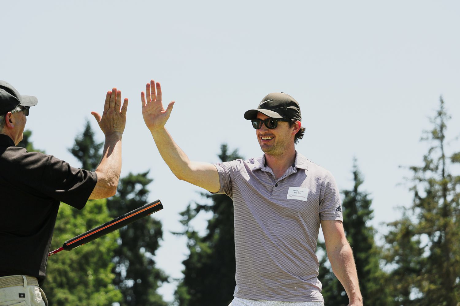 Two men high-fiving on a golf course with tall trees and blue sky in the background.