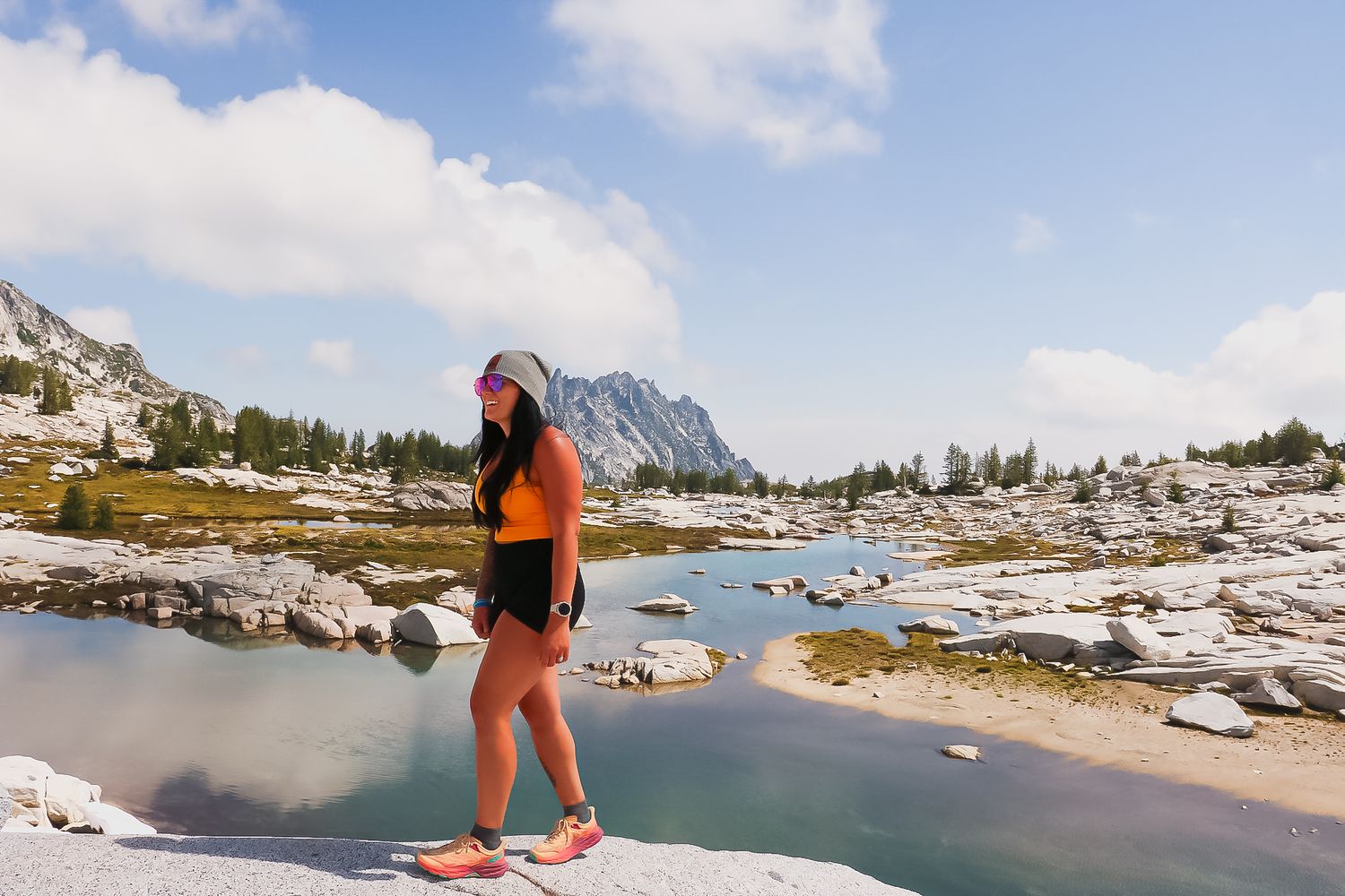A woman hiker stands by a serene alpine lake surrounded by rocky terrain and mountains under a partly cloudy sky.