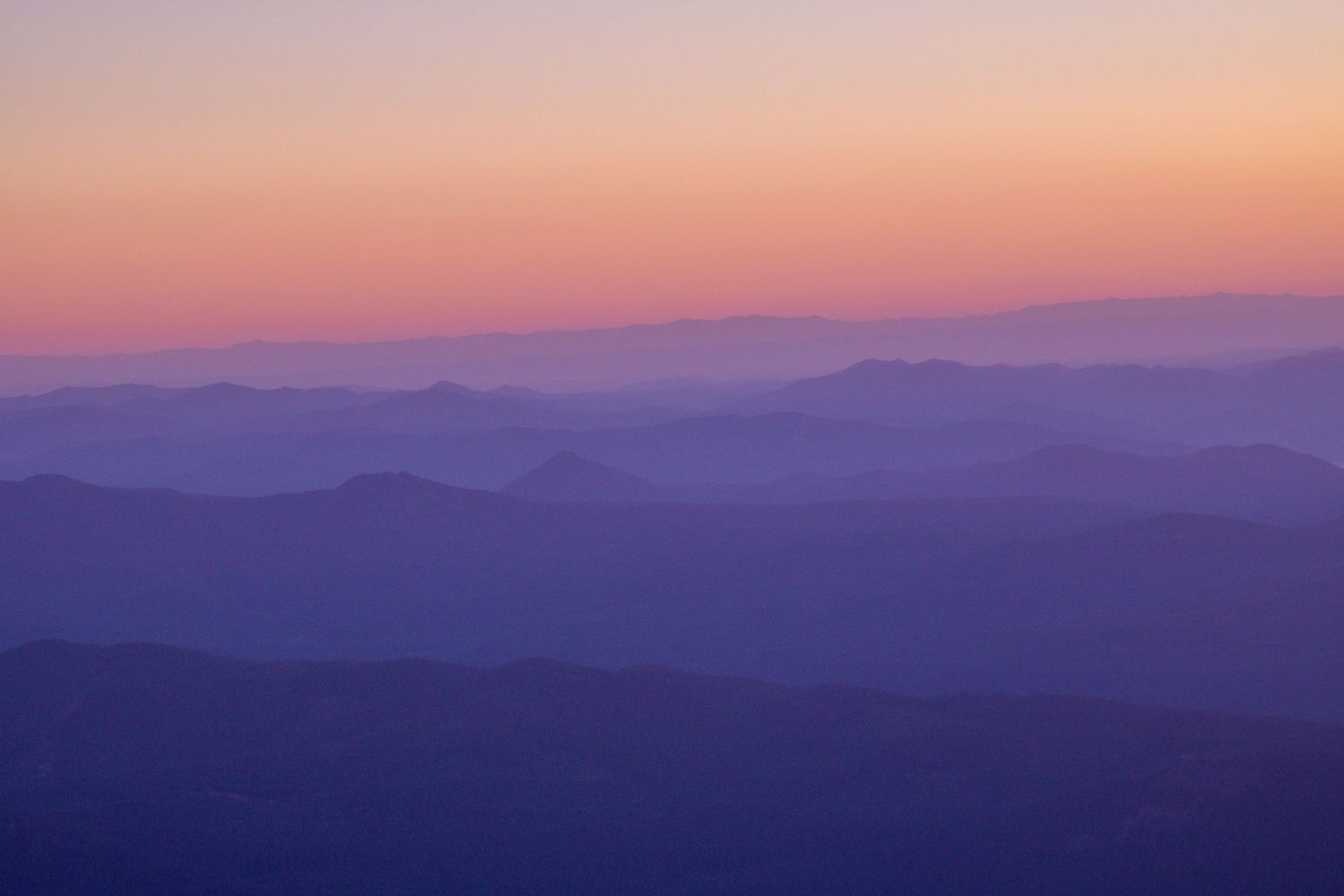 Layered mountain silhouettes fade into a stunning purple and orange gradient sunset sky over a vast wilderness landscape.