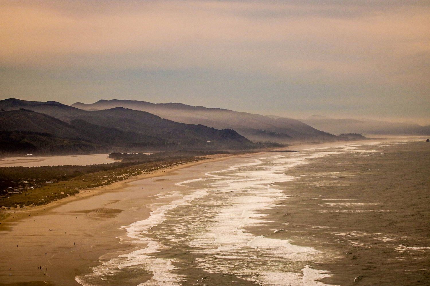 Aerial view of a long sandy coastline with rolling waves and misty mountains at dusk.