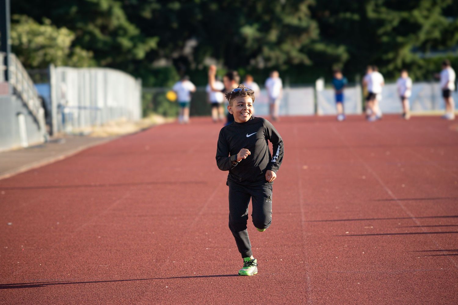 Child in black Nike outfit jogging alone on a red track in sunlight.