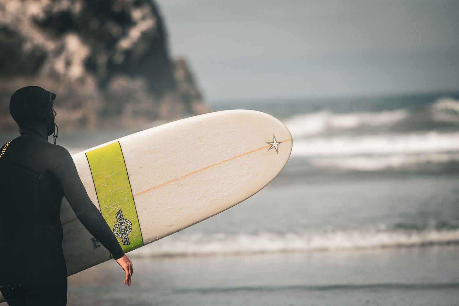 Surfer in black wetsuit holding a white and yellow longboard on a rocky beach with ocean waves.