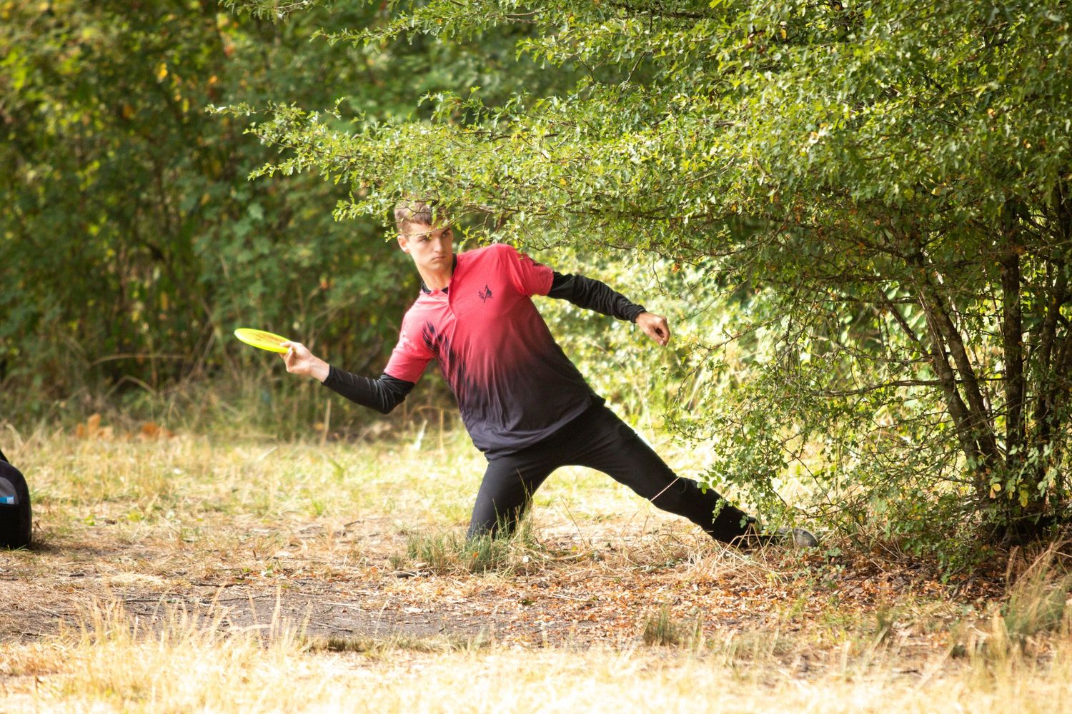 Young man in red shirt throwing a yellow disc golf frisbee near dense trees outdoors.