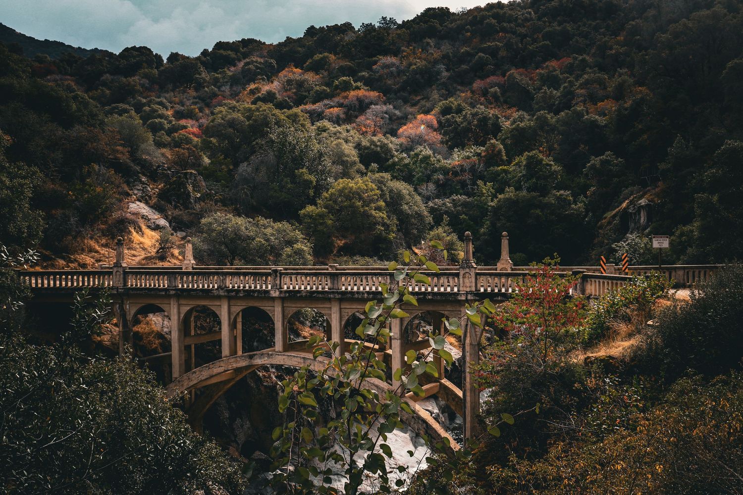 A historic stone arch bridge spans a rocky canyon surrounded by dense autumn foliage on a hillside.