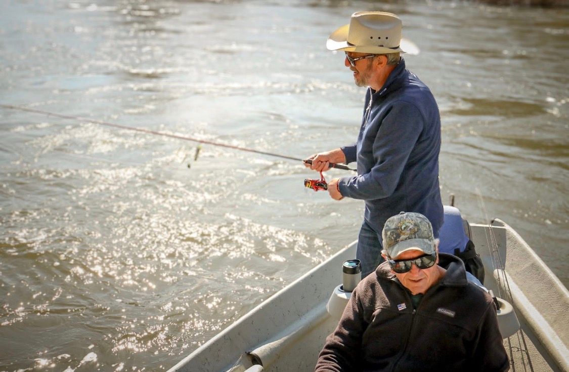Two men fish from a small boat on a sparkling river.
