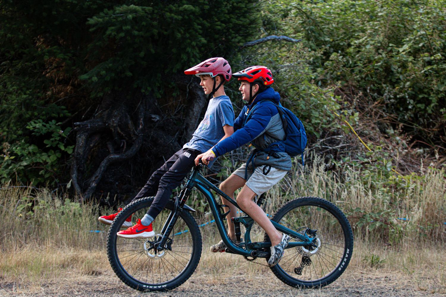 Two children wearing helmets riding together on a mountain bike along a dirt trail.