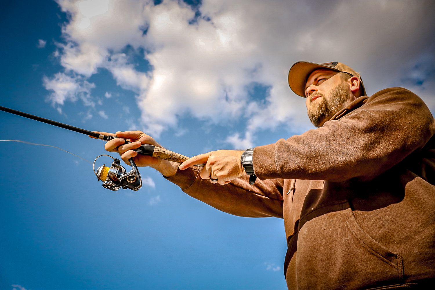 A bearded man in a brown jacket casts a fishing rod against a bright blue cloudy sky.
