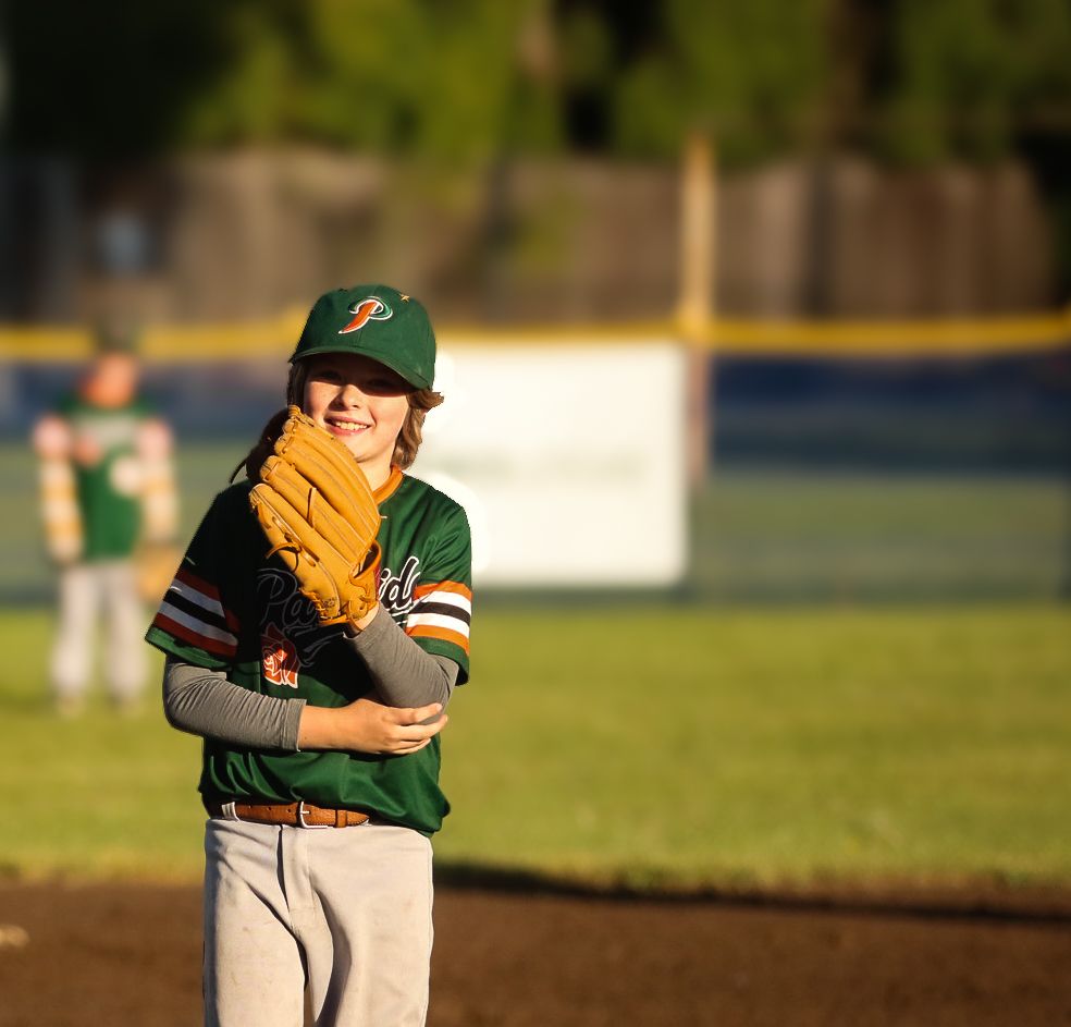 Young baseball player in green uniform and cap smiling on field, holding glove during a sunny game.