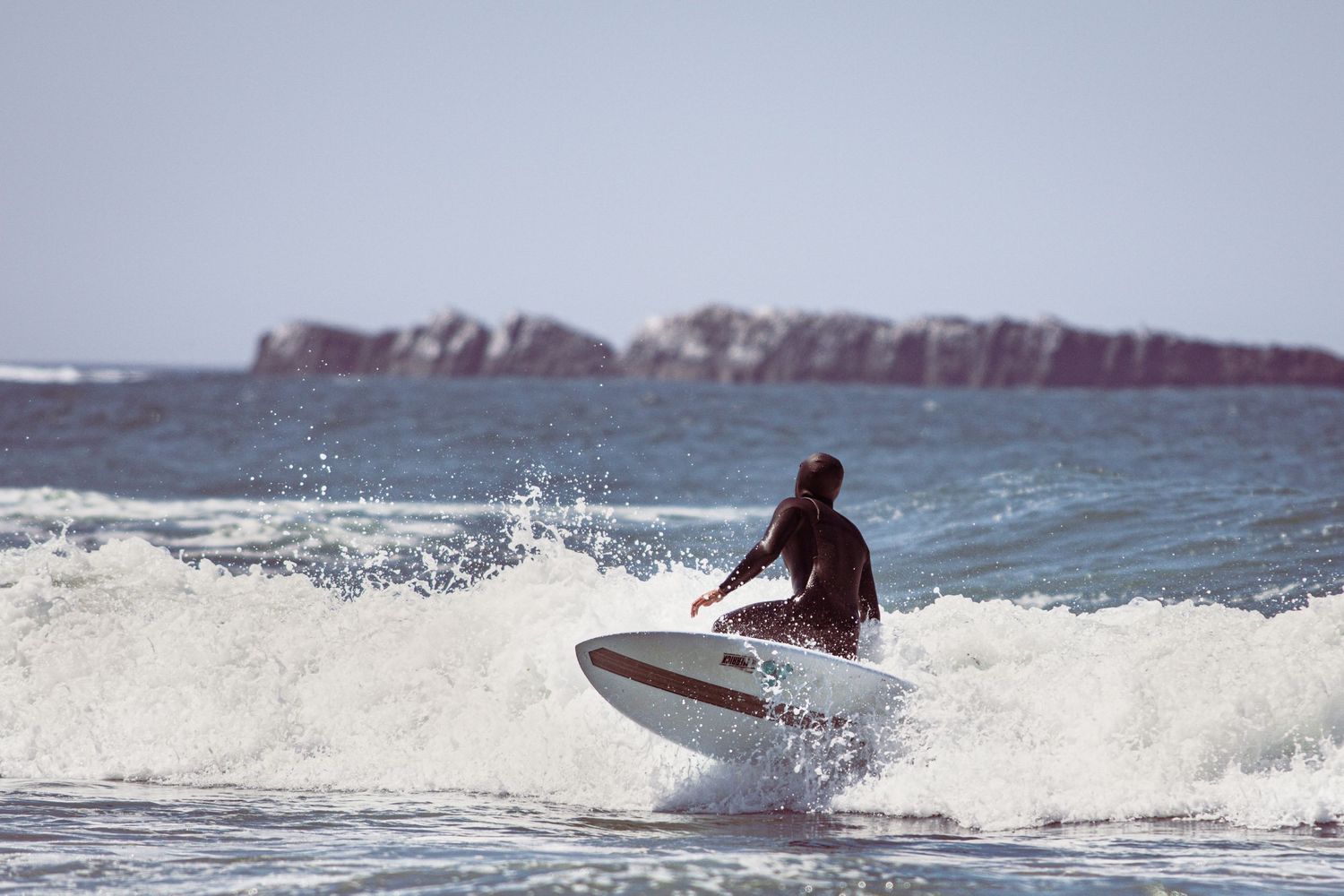 A surfer in a wetsuit rides a white wave on a bright sunny day near rocky ocean outcrops.