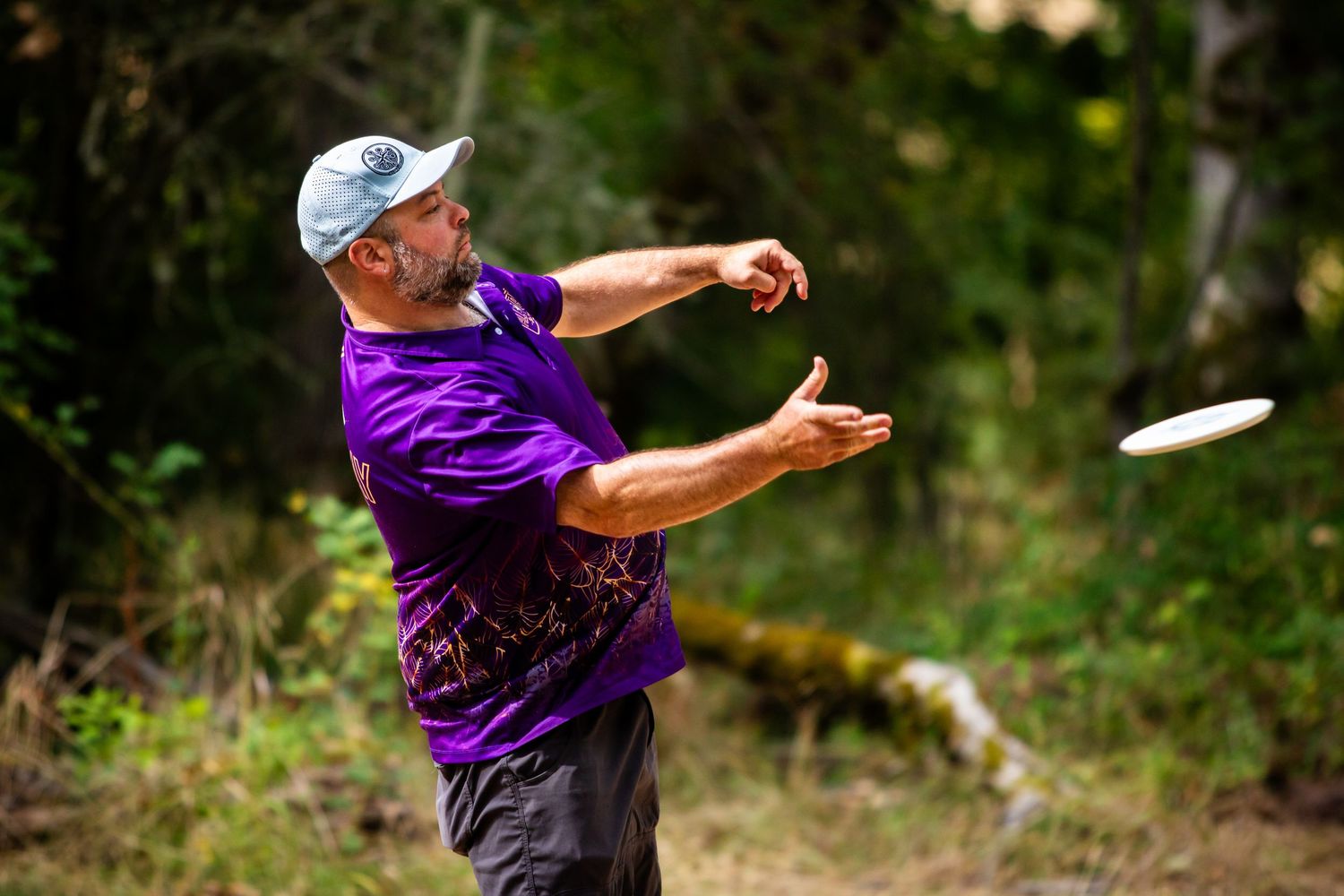 Older man in purple shirt releasing a white frisbee while playing disc golf in a forest.
