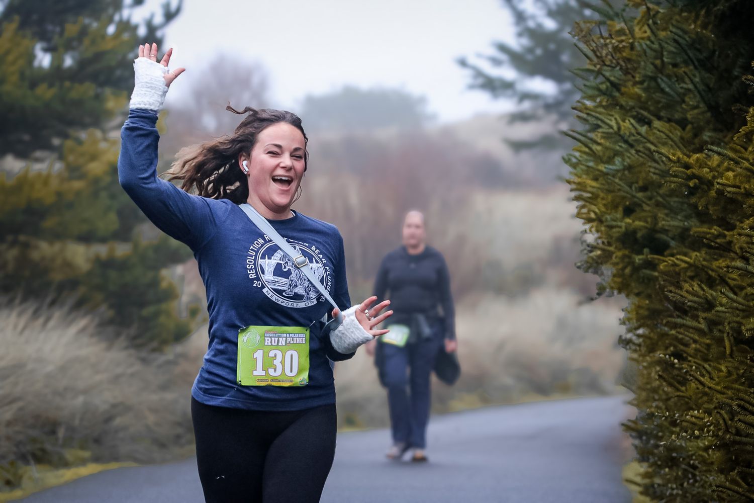A cheerful woman with bib number 130 waves while running a race near evergreen trees.