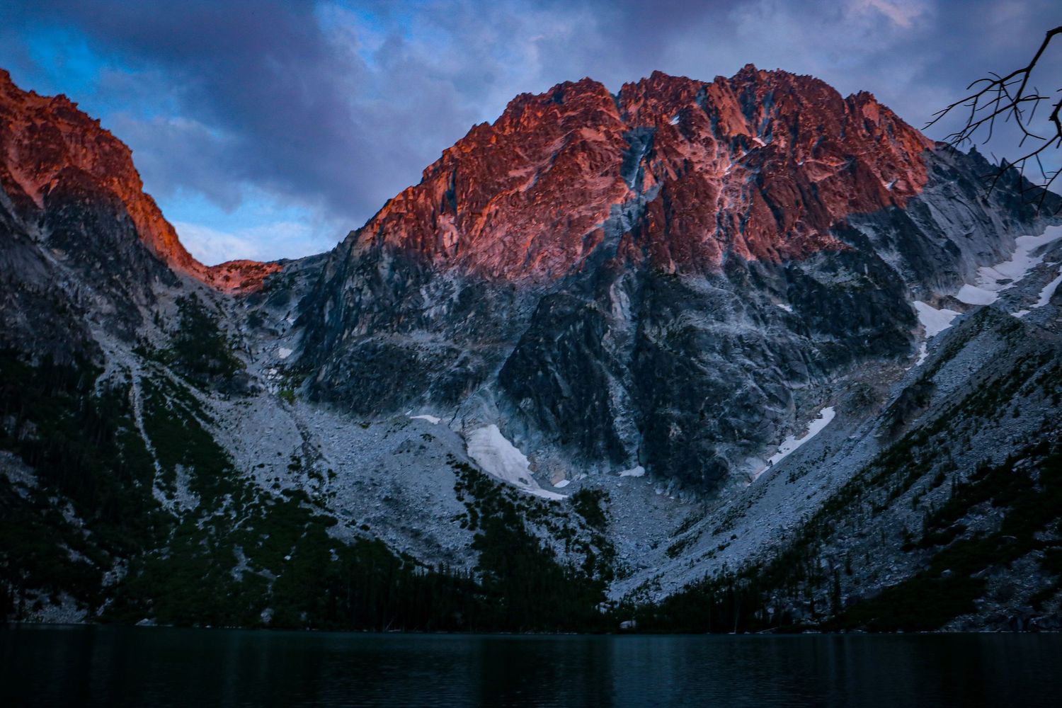 Fiery red alpenglow illuminates rugged snow-dusted mountain peaks above a dark alpine lake at sunset.