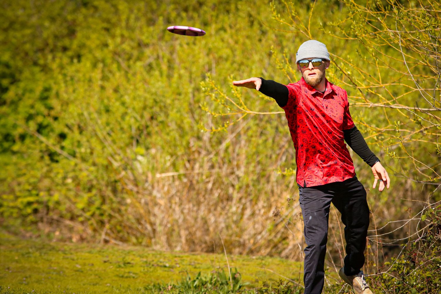 Woman in red jacket throwing a disc golf frisbee on a lush green course.