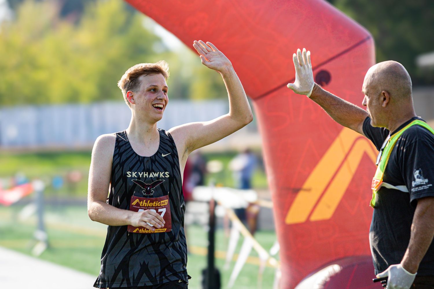Young runner in Skyhawks jersey high-fiving a volunteer at a race finish line outdoors.