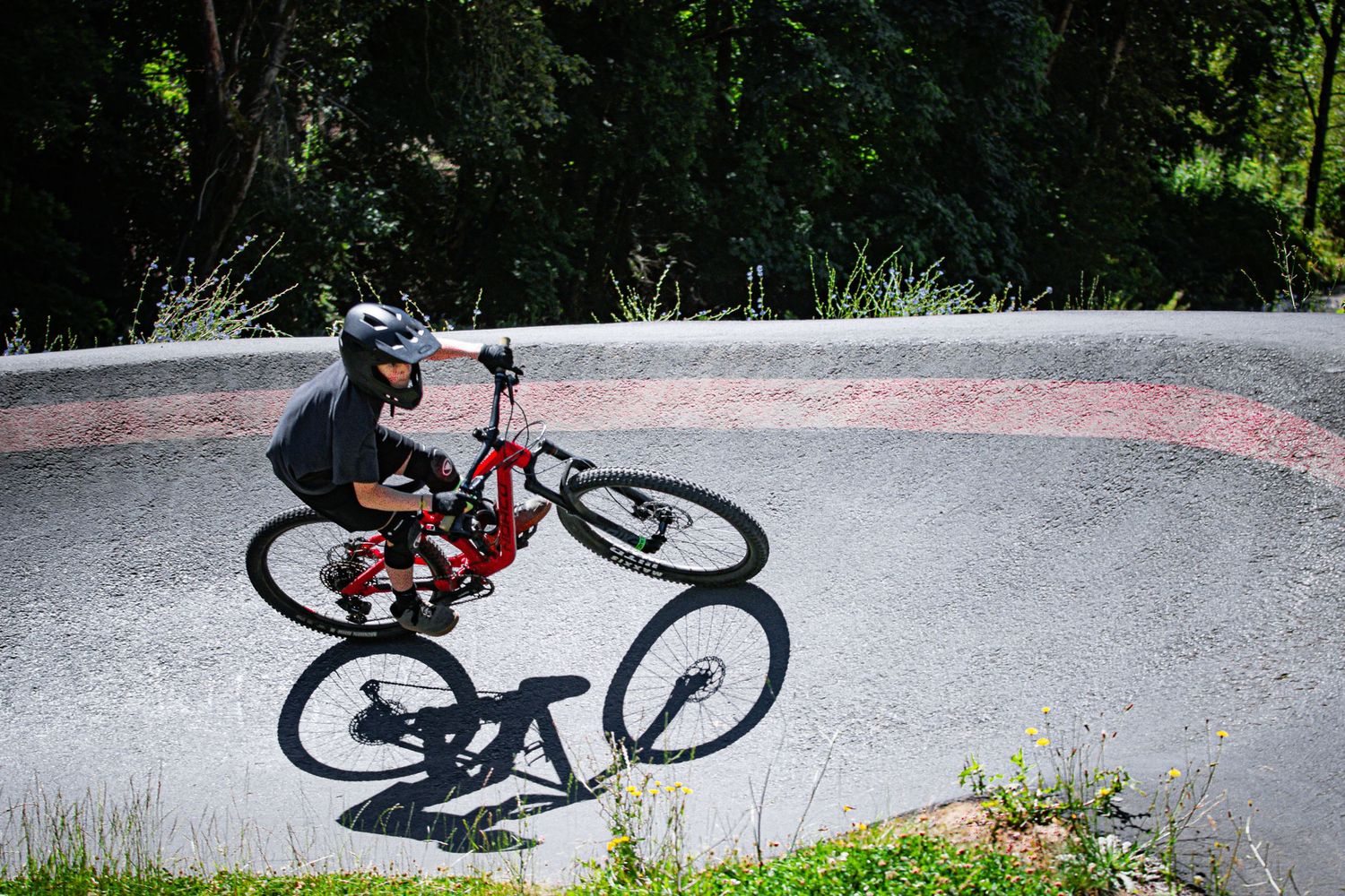 A helmeted rider performs a wheelie on a red mountain bike at a concrete pump track park.