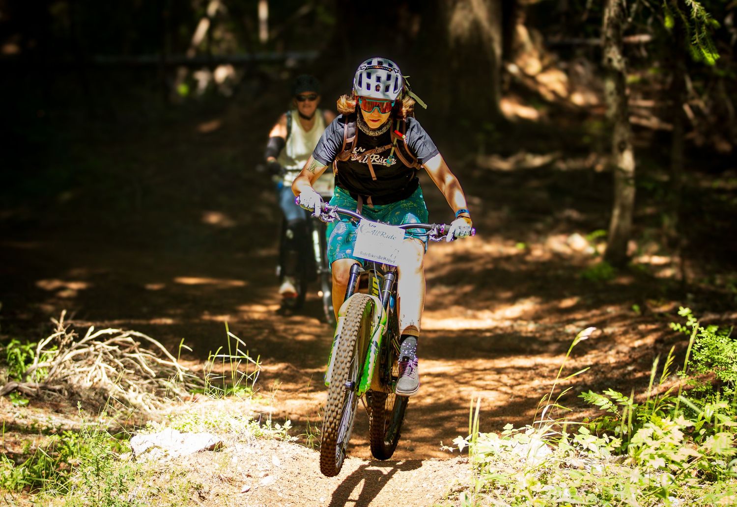 Mountain biker riding downhill on forest trail with another cyclist following behind.