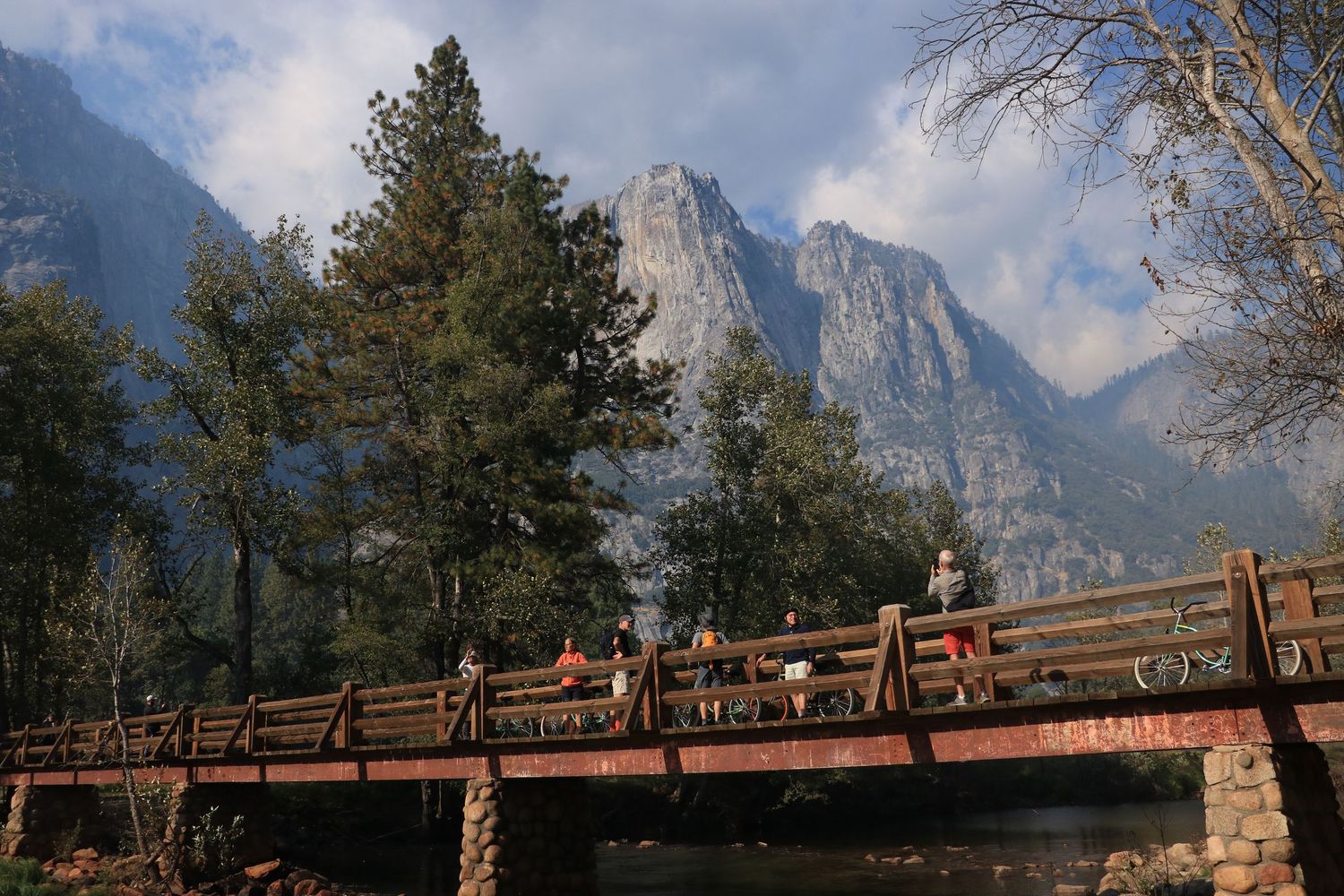Wooden bridge over a river with hikers and dramatic mountain peaks rising behind pine trees.