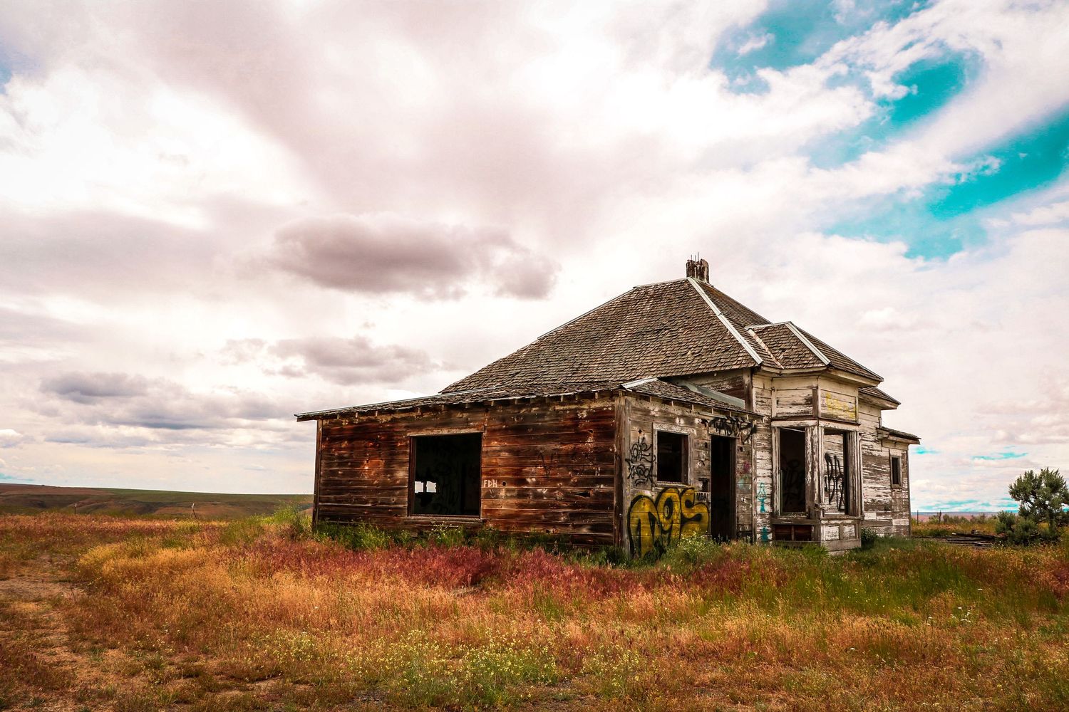 Abandoned weathered wooden farmhouse with graffiti standing alone on an open prairie under cloudy skies.