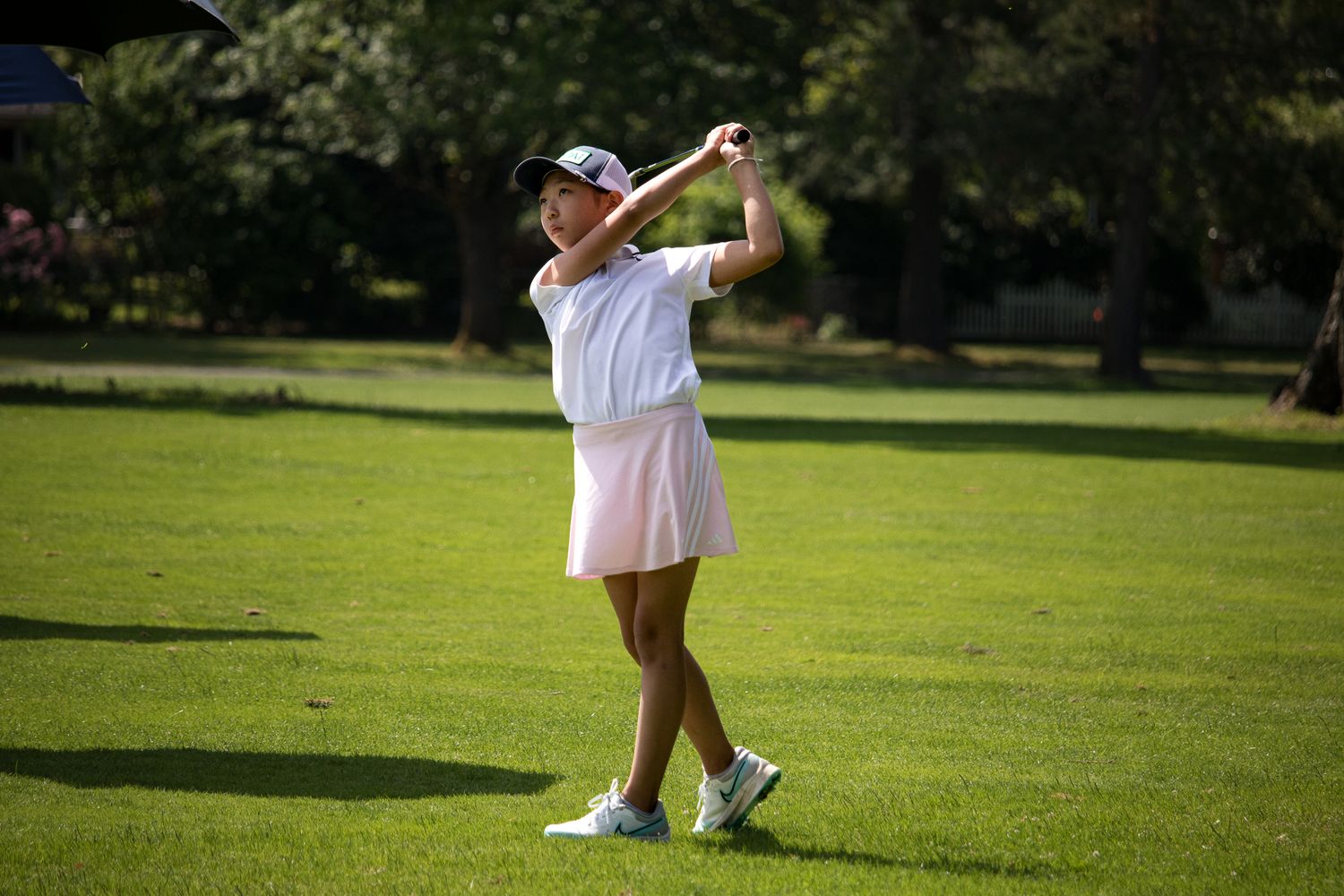 Young female golfer in pink skirt swinging a club on a sunny green fairway.
