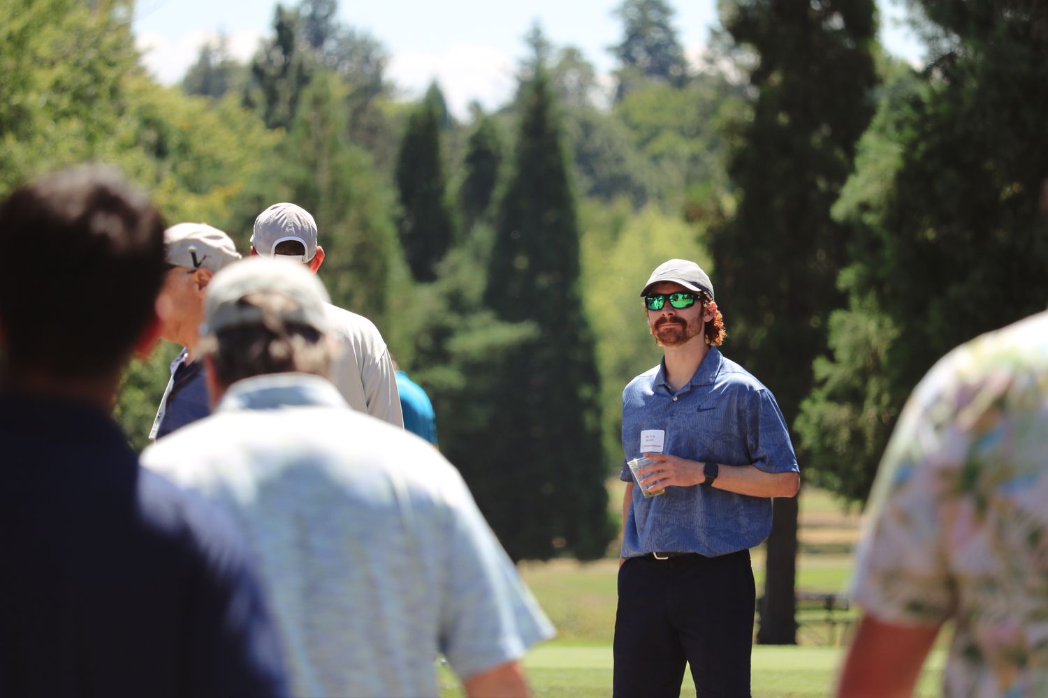 A man in a blue shirt and sunglasses addresses a group of golfers on a sunny course.