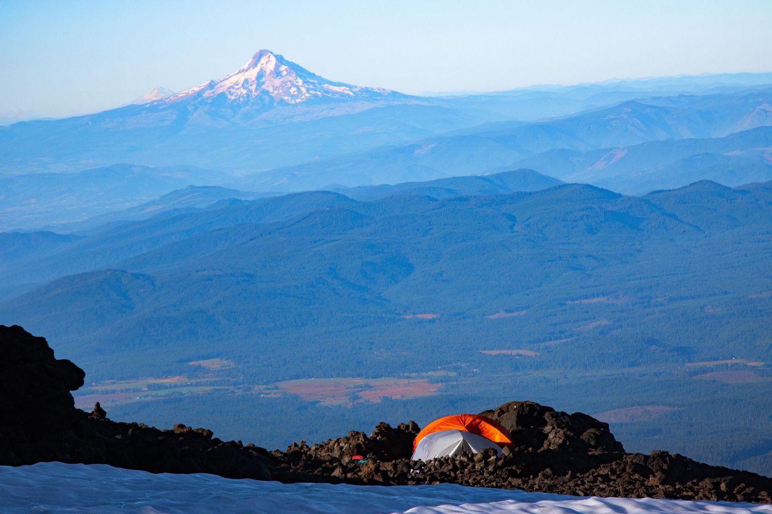 A small orange tent sits on a snowy ridge with a distant snow-capped volcanic peak visible through blue mountain haze.