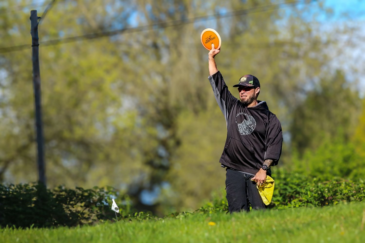 Man in black hoodie throwing an orange disc golf frisbee on a grassy course.