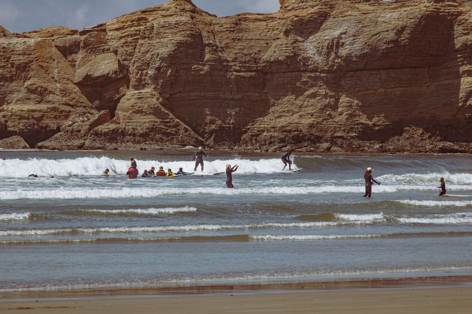 Surfers and swimmers enjoy waves near dramatic golden sandstone cliffs along a scenic beach.