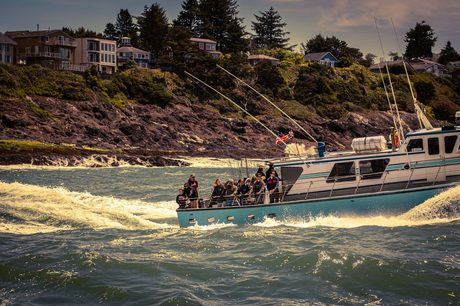 A fishing charter boat cruises along a rocky coastline with passengers on board during golden hour.