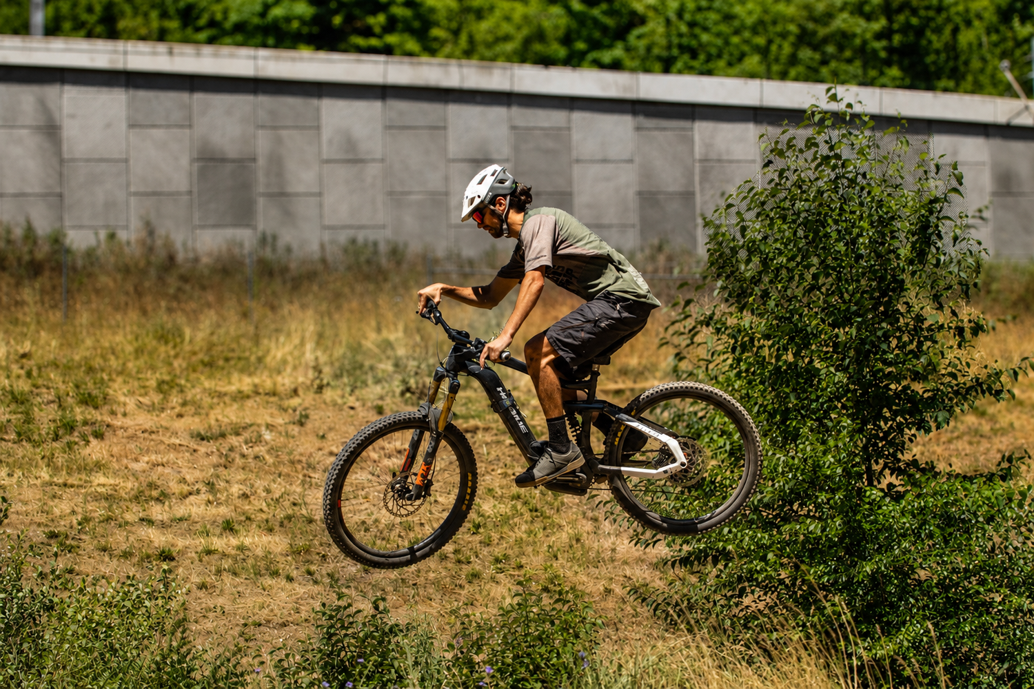A mountain biker in a white helmet rides through dry grass near a concrete wall on a sunny day.