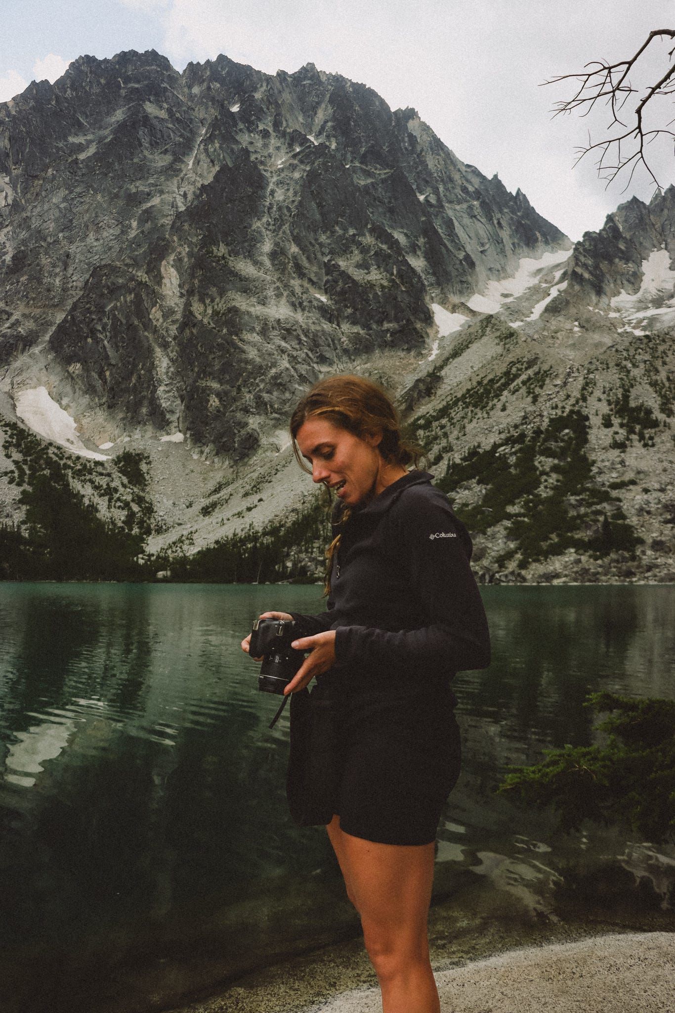 A woman in black jacket holds a camera by a alpine lake with dramatic rocky mountain peaks.