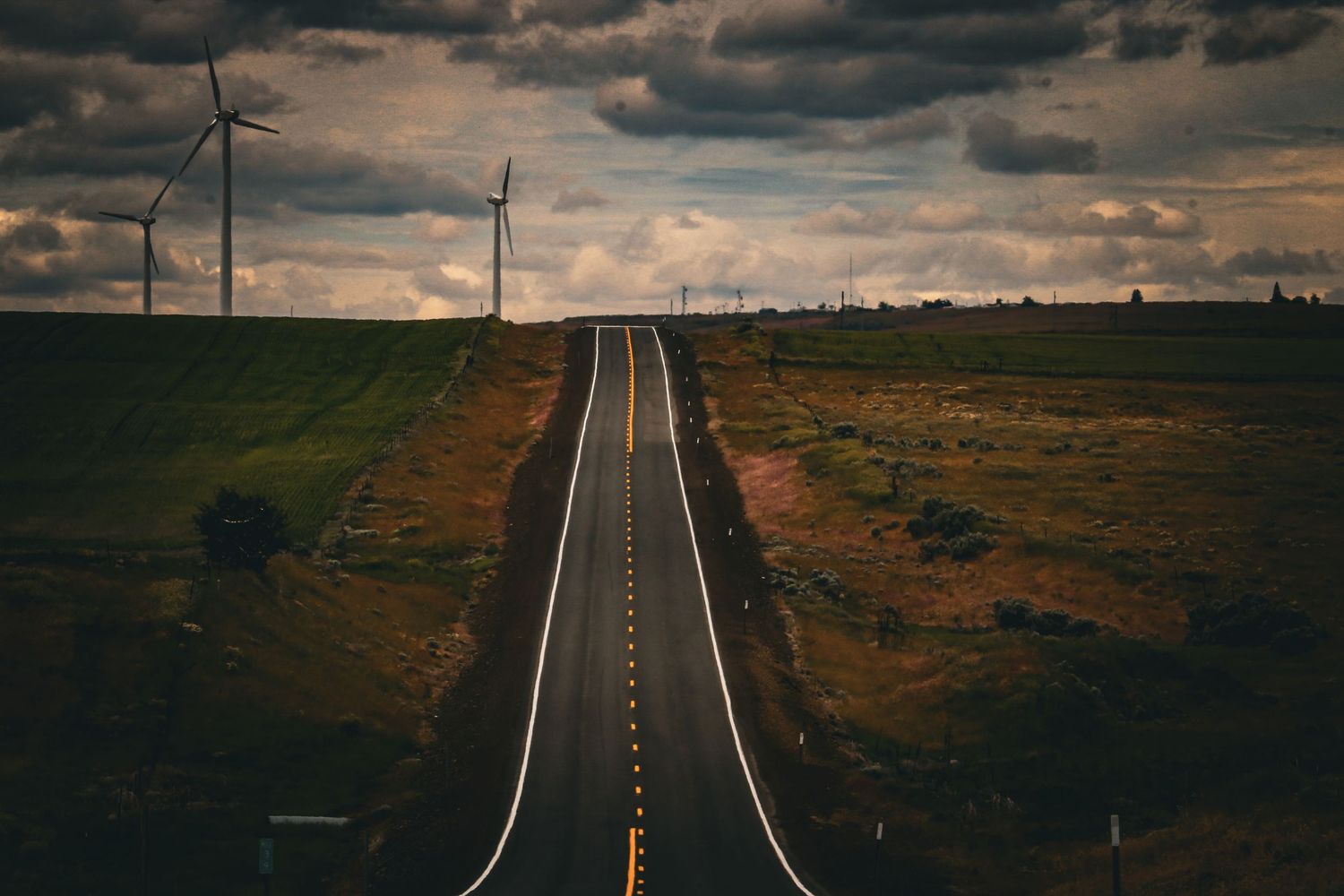 Straight highway stretching to the horizon flanked by wind turbines under dramatic storm clouds.